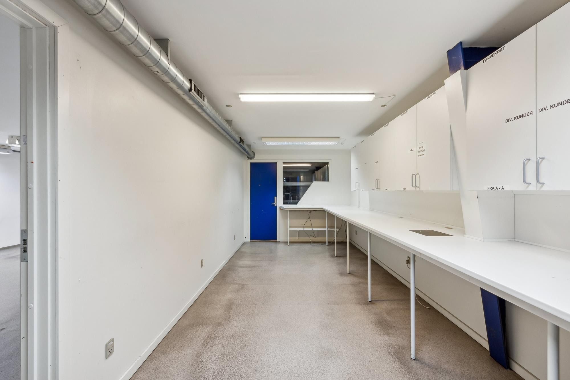 Et tomt kontor med hvide vægge og loft, højt til loftet og gulvtæppe, slanke arbejdsborde, slanke højre hængeskabe og en