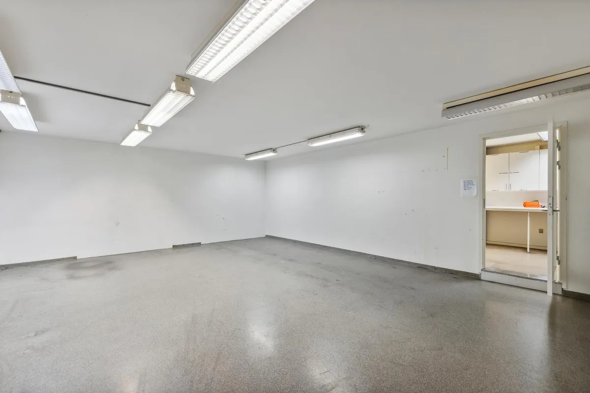 Et ubenyttet lagerrum med to store lysekroner og en åben dør, der fører til et mindre rum med et bord og høje skabe