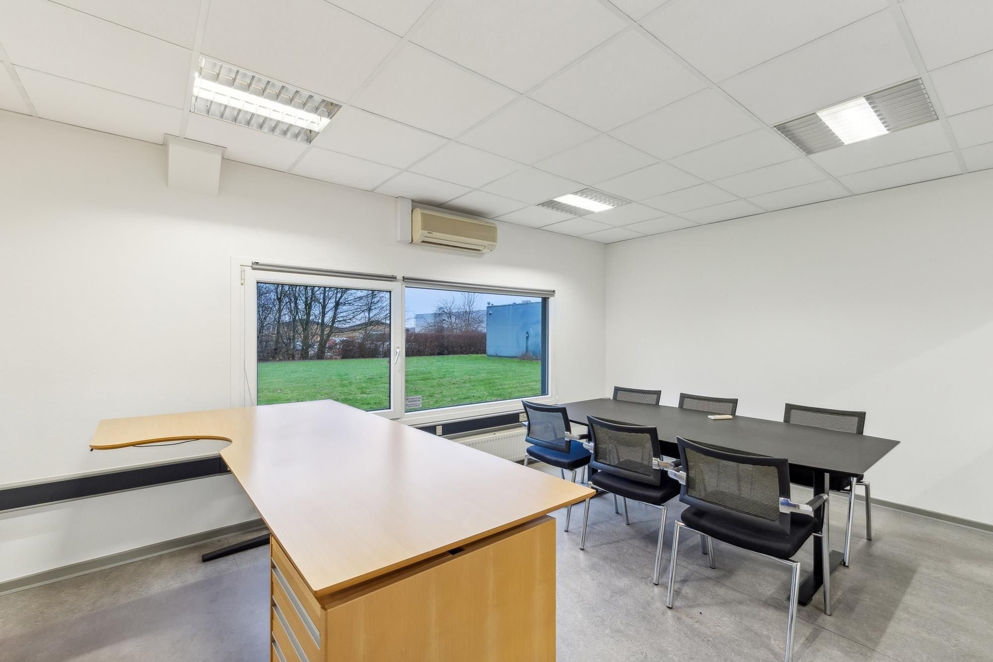 Et stort kontor med hvide vægge og loft samt gulvtæppe og vinduer med udsigt til græsplæne og bygninger i baggrunden