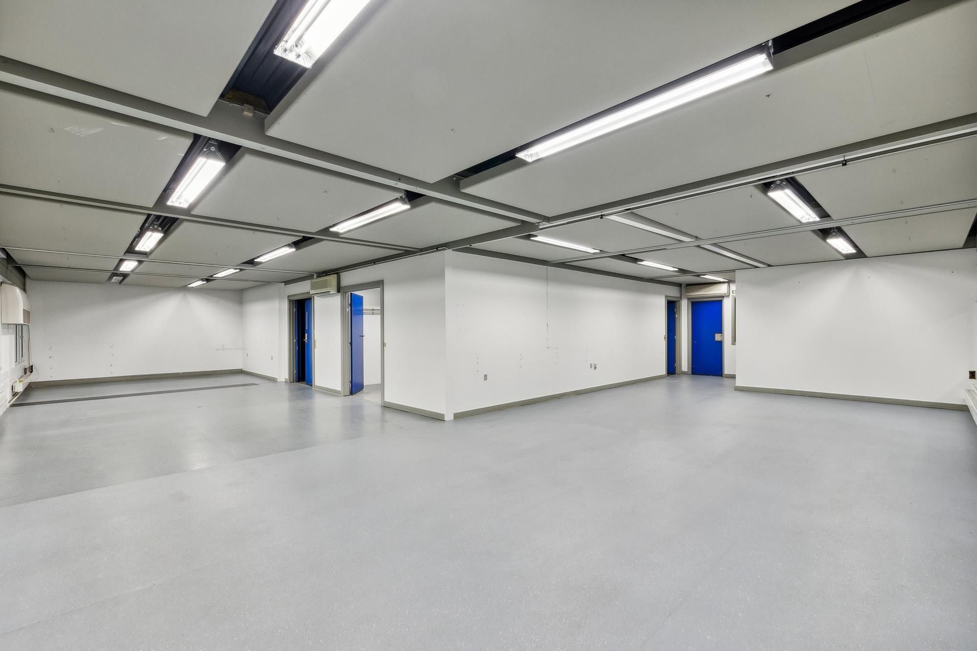 En stor, ubenyttet lagerhal med hvide vægge og gulve samt to blå indgangsdøre og lyse plafonder med lysearmaturer
