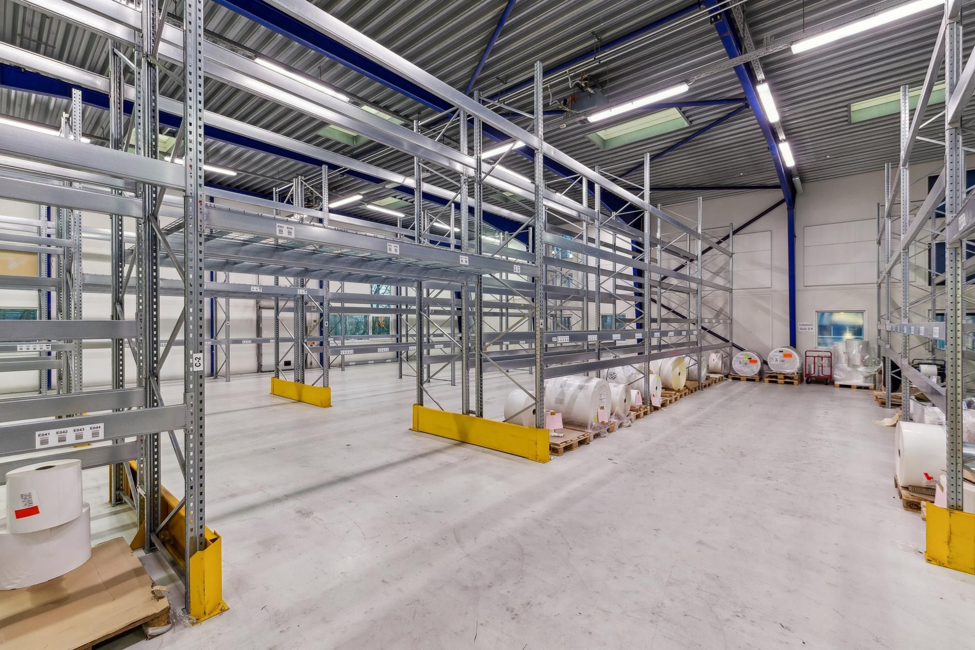 Lager med mange høje metalreoler og gulve. Gulvet er glat og renset, og der er flere ruller og pakker pakket på palle.