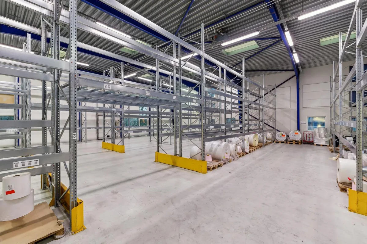 Lager med mange høje metalreoler og gulve. Gulvet er glat og renset, og der er flere ruller og pakker pakket på palle.