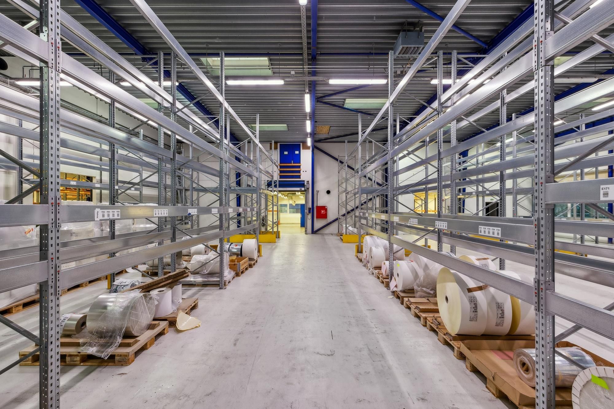 Lager med mange metalhylder og pallehylderne fyldt med ruller, folie og karton. En dør er åben, og der er gulvet er hvidt.