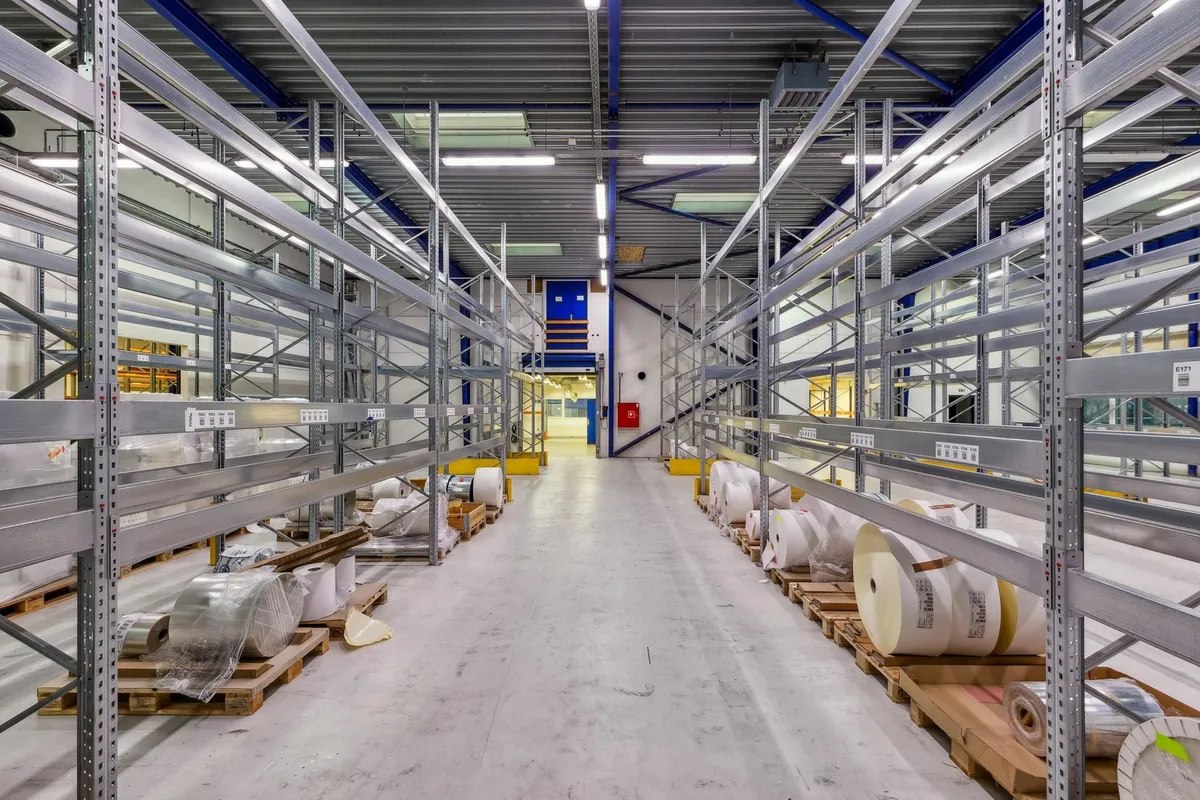 Lager med mange metalhylder og pallehylderne fyldt med ruller, folie og karton. En dør er åben, og der er gulvet er hvidt.