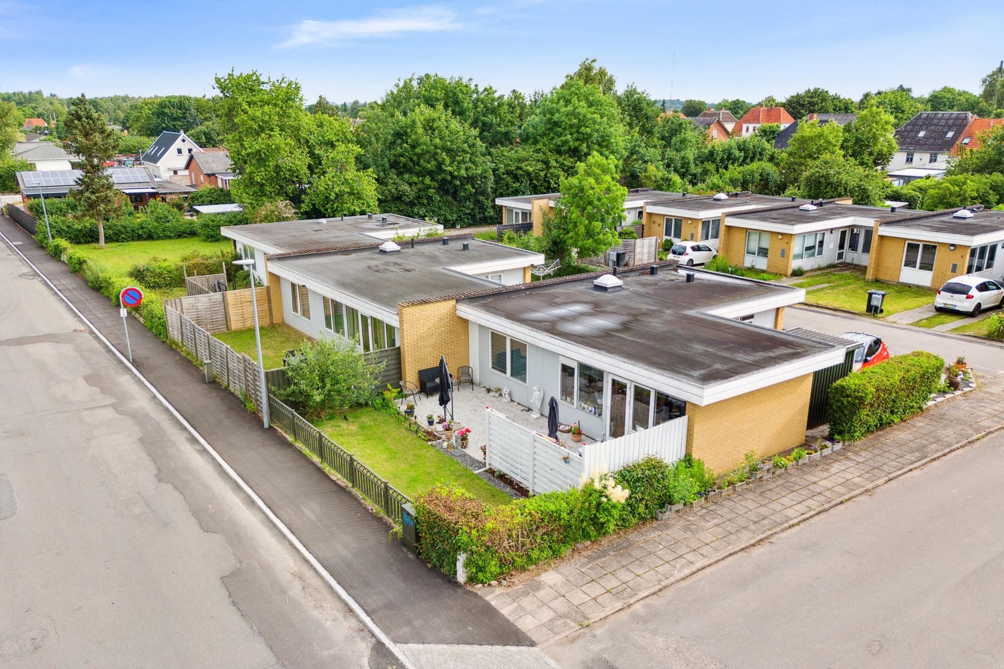 16 moderne rækkehuse med hver deres have og solceller på taget, omgivende grønne områder og lyseblå himmel.