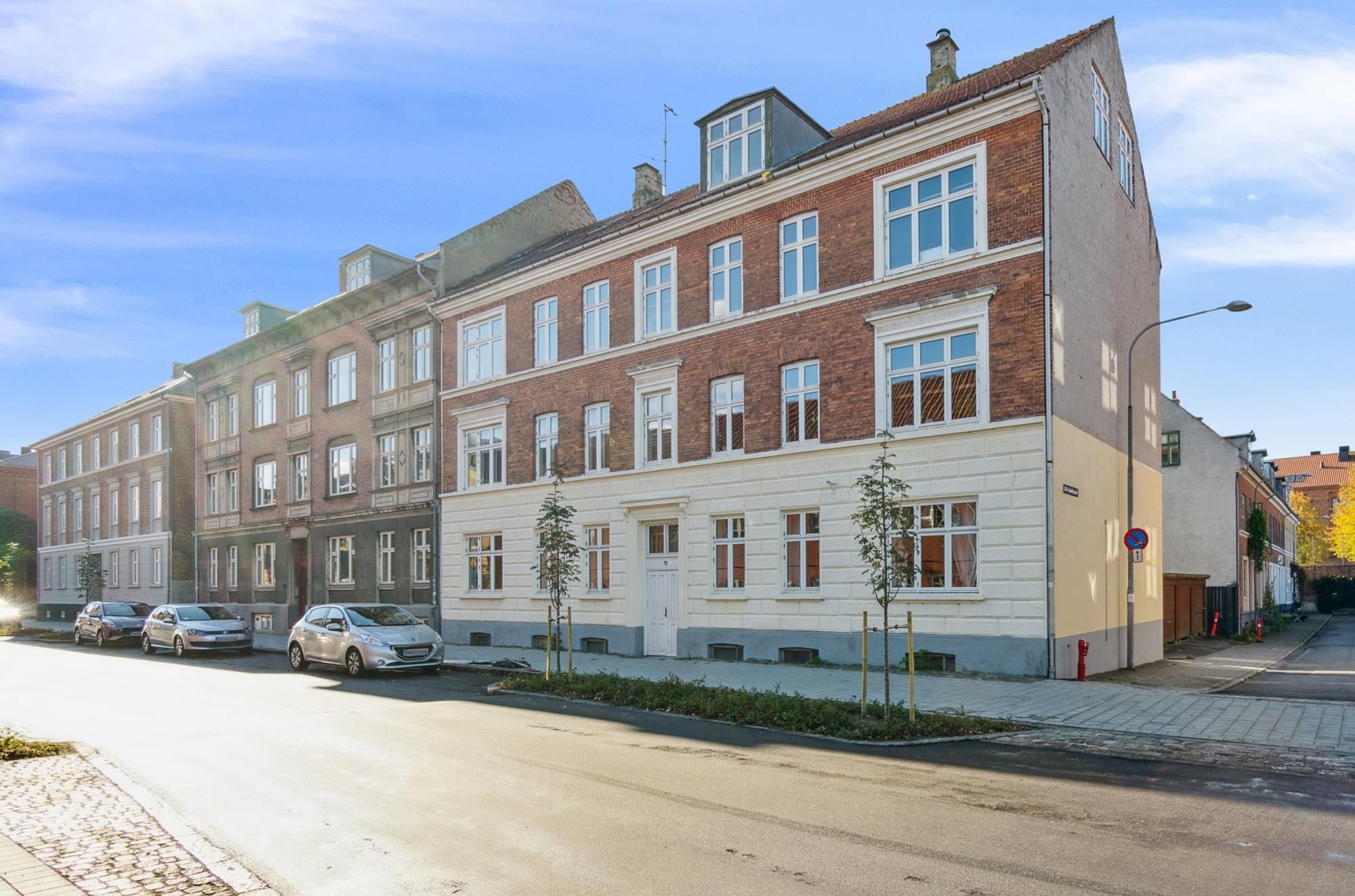 En smuk ejendom med røde mursten og hvide detaljer står på en gade i en lille by med tre parkerede biler og en søjle