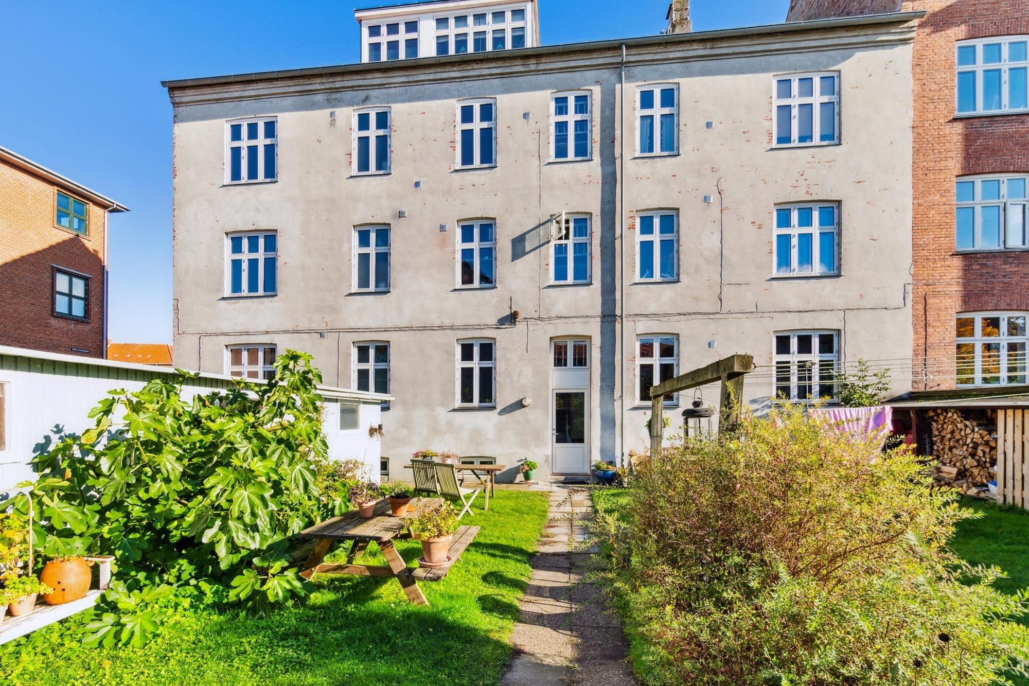 Småhusejendom med 7 boliger og haveområde. I baghaven er der en picnicbord, række med plantepotter og et lysanlæg.