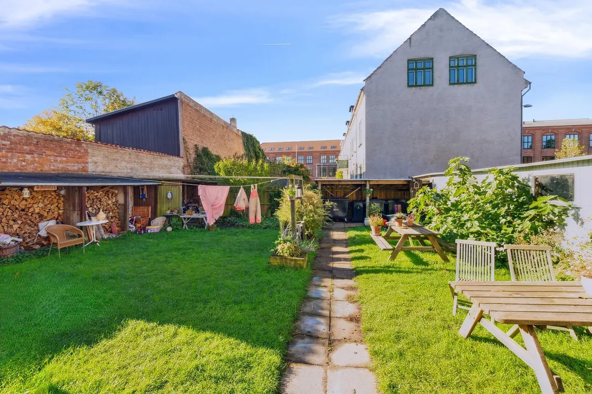 Småhusejendom med smuk have og lyse blomster, træborde og stole samt en opbevaringsbygning med træstamme og klædtøj