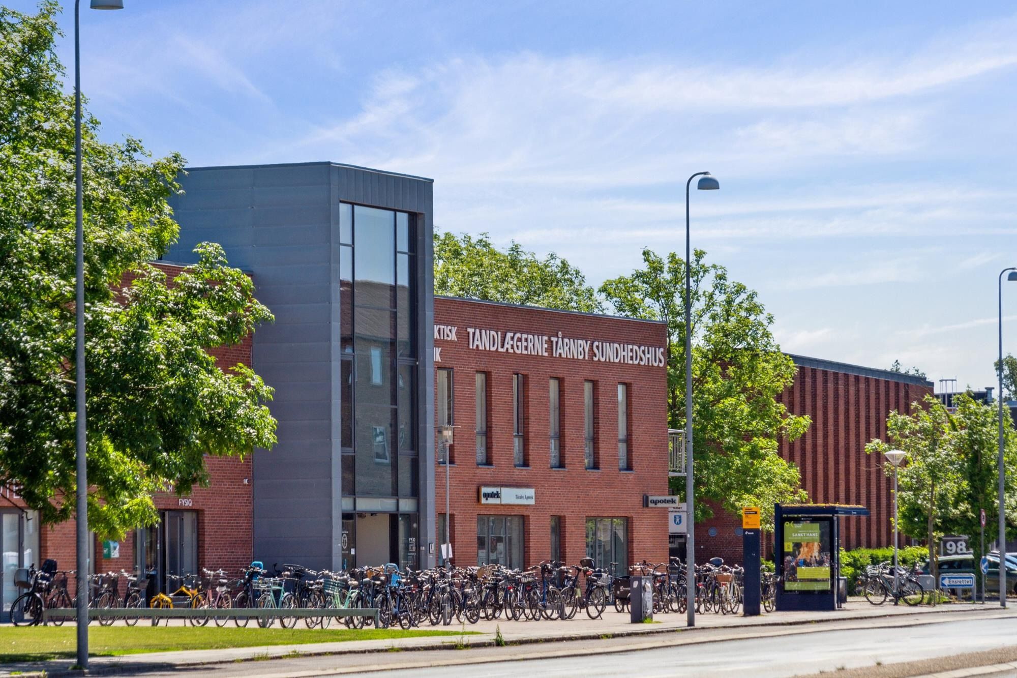 Bygning med røde mursten og glasvinduer, skilte med teksten "Tandlægerne Tårnby Sundhedshus" og "optik", mange cykler