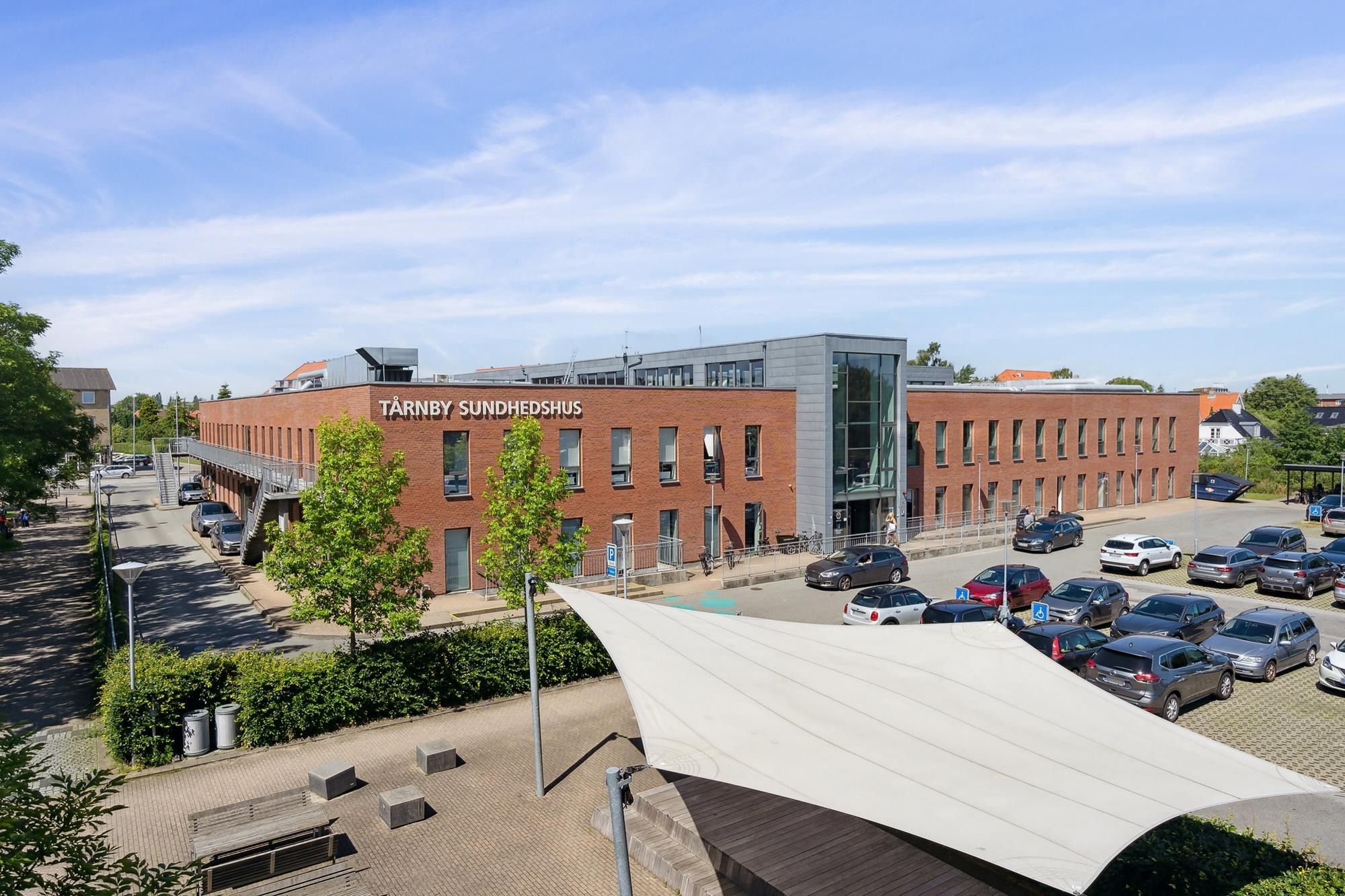 Tårnby Sundhedshus med parkeringsplads og udsigt over grønne træer og buske og en hvid skyggeskærm i forgrunden