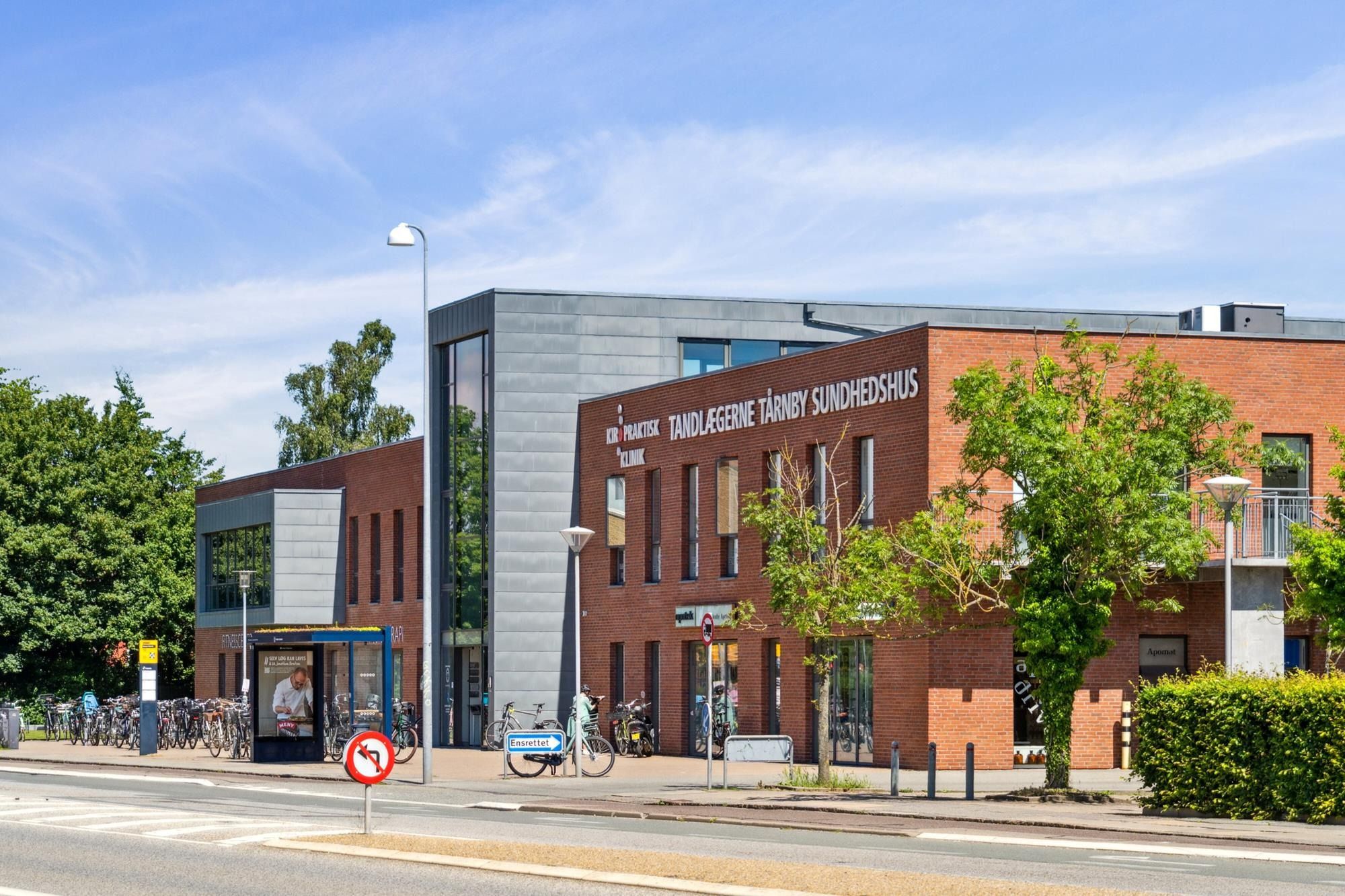 Tårnby Sundhedshus med busstoppested, cykelparkering og grønne træer på en solrig dag