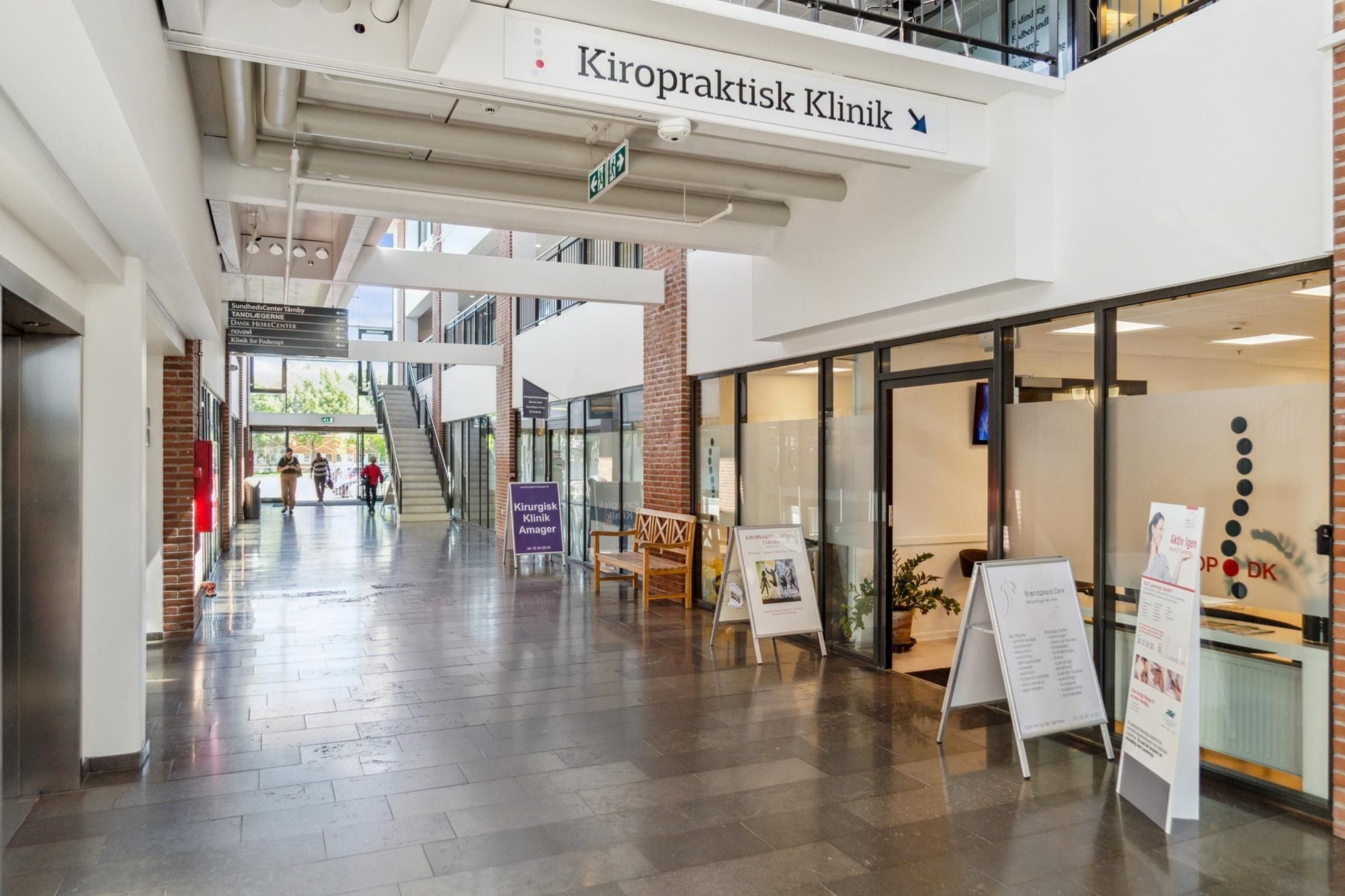 Indgående gangvej med skiltet Kiropraktisk Klinik. Folk går i forgrunden, og der er en busk og stol i nærheden af vinduet.