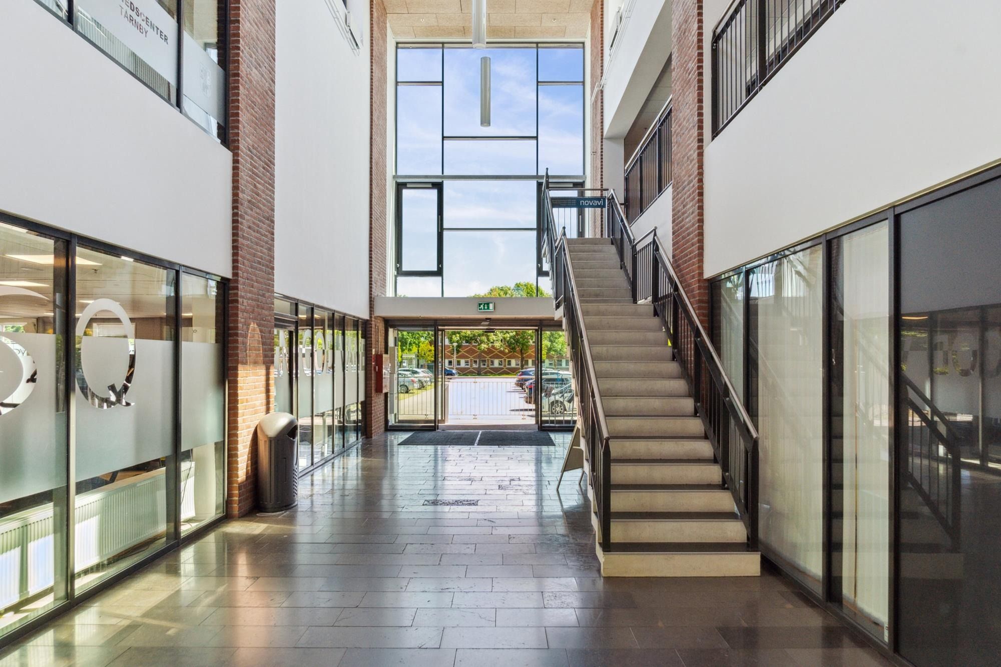 Hallen i fuld udlejet ejendom med store vinduer og lysegule gulvfliser og skrå trappe med mørke håndtag