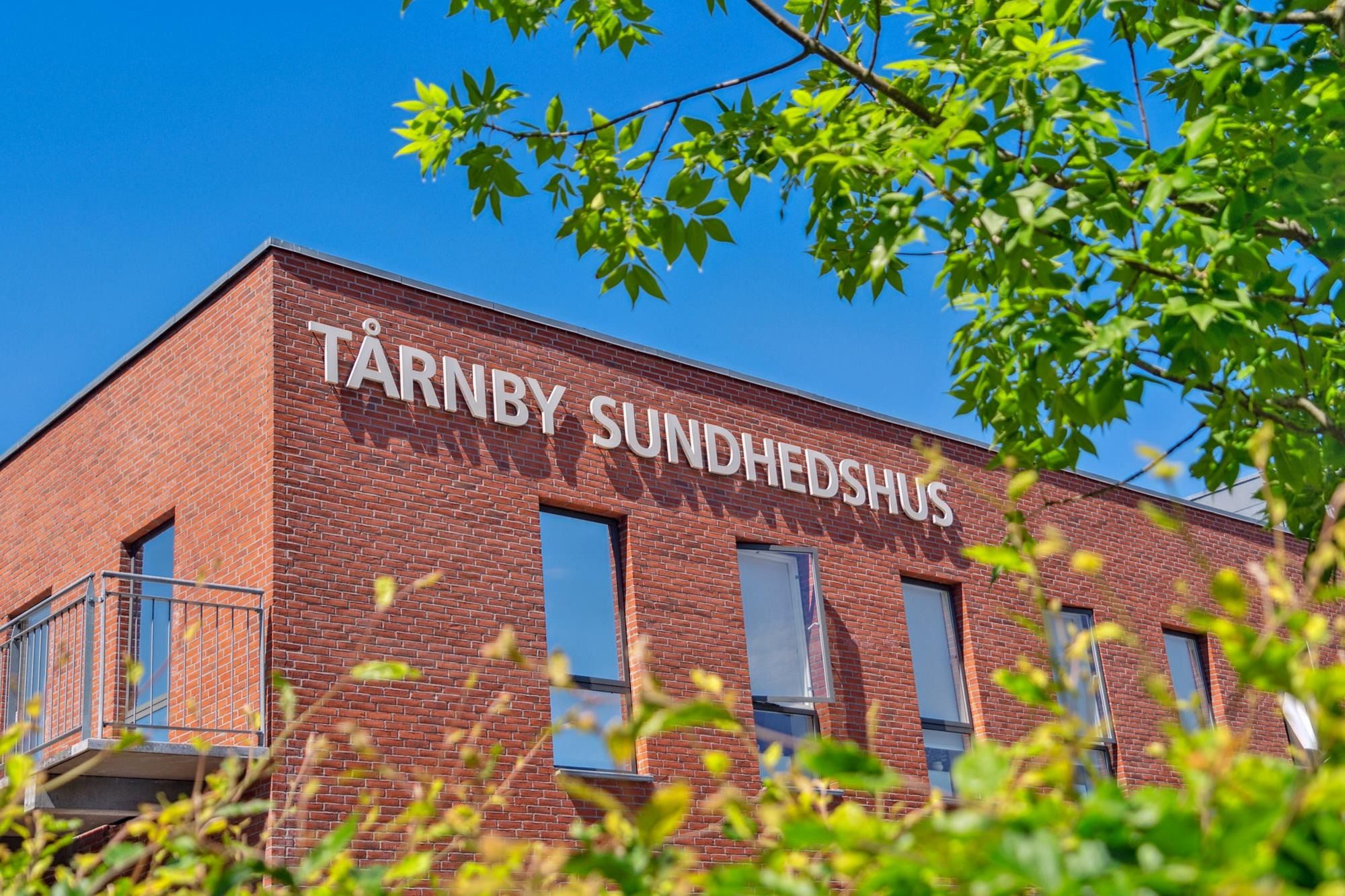 Tårnby Sundhedshus bygning med skilte, røde mursten og glasvinduer under en lyseblå himmel med grønne løv