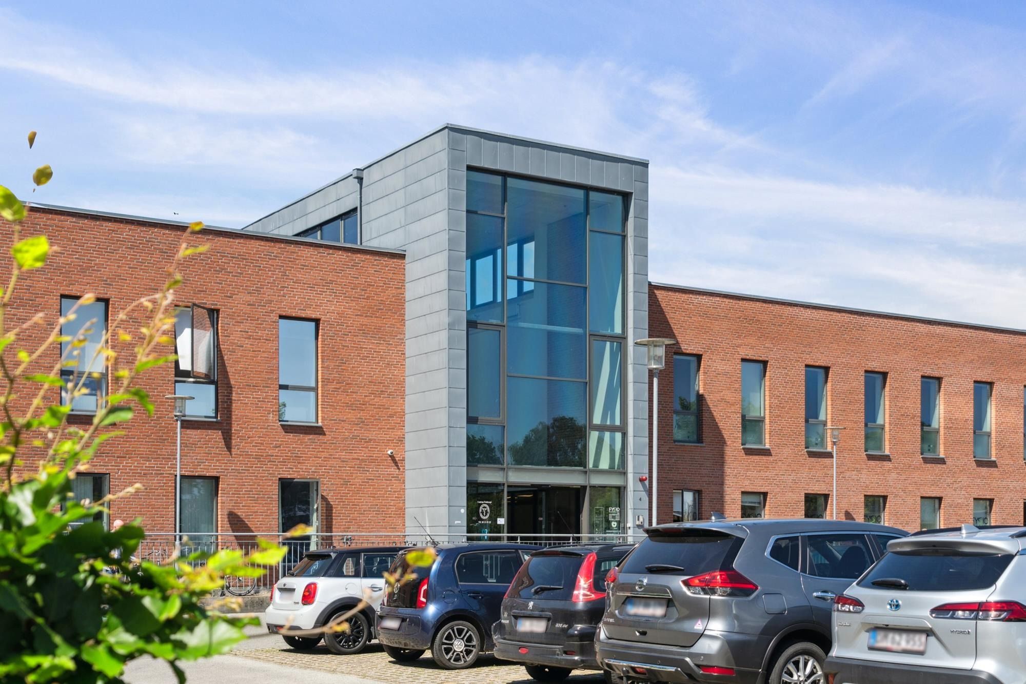 Moderne bygning med store glasruder og lyse facader er omgivet af flere parkerede biler og grønne planter