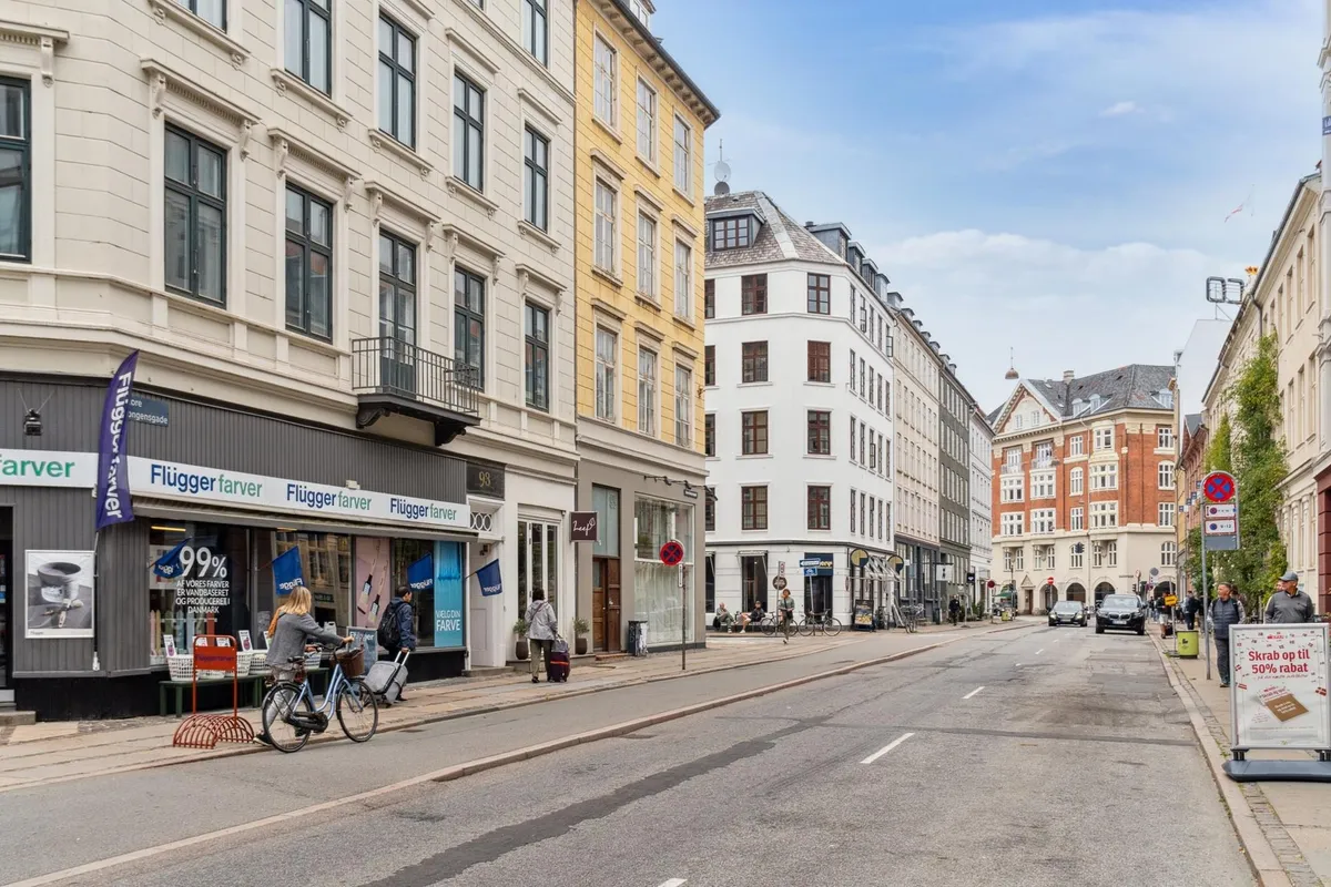Store Kongensgade med en cykelkørende og folk, der går og står, sammen med butikker, biler og husene med mange vinduer