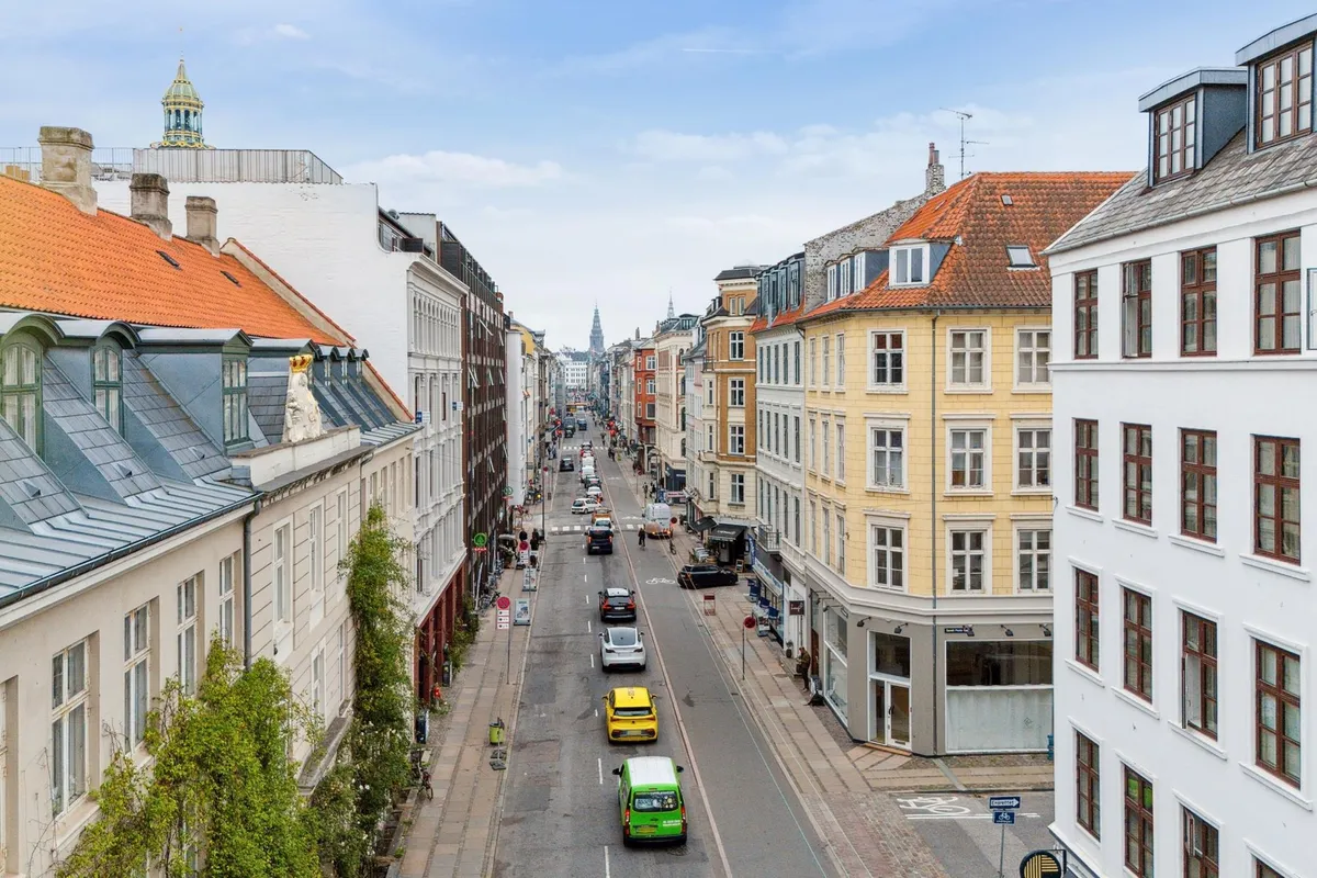 Biler kører gennem Københavns gade, forbi bygninger med røde tag, hvor folk går og cykler