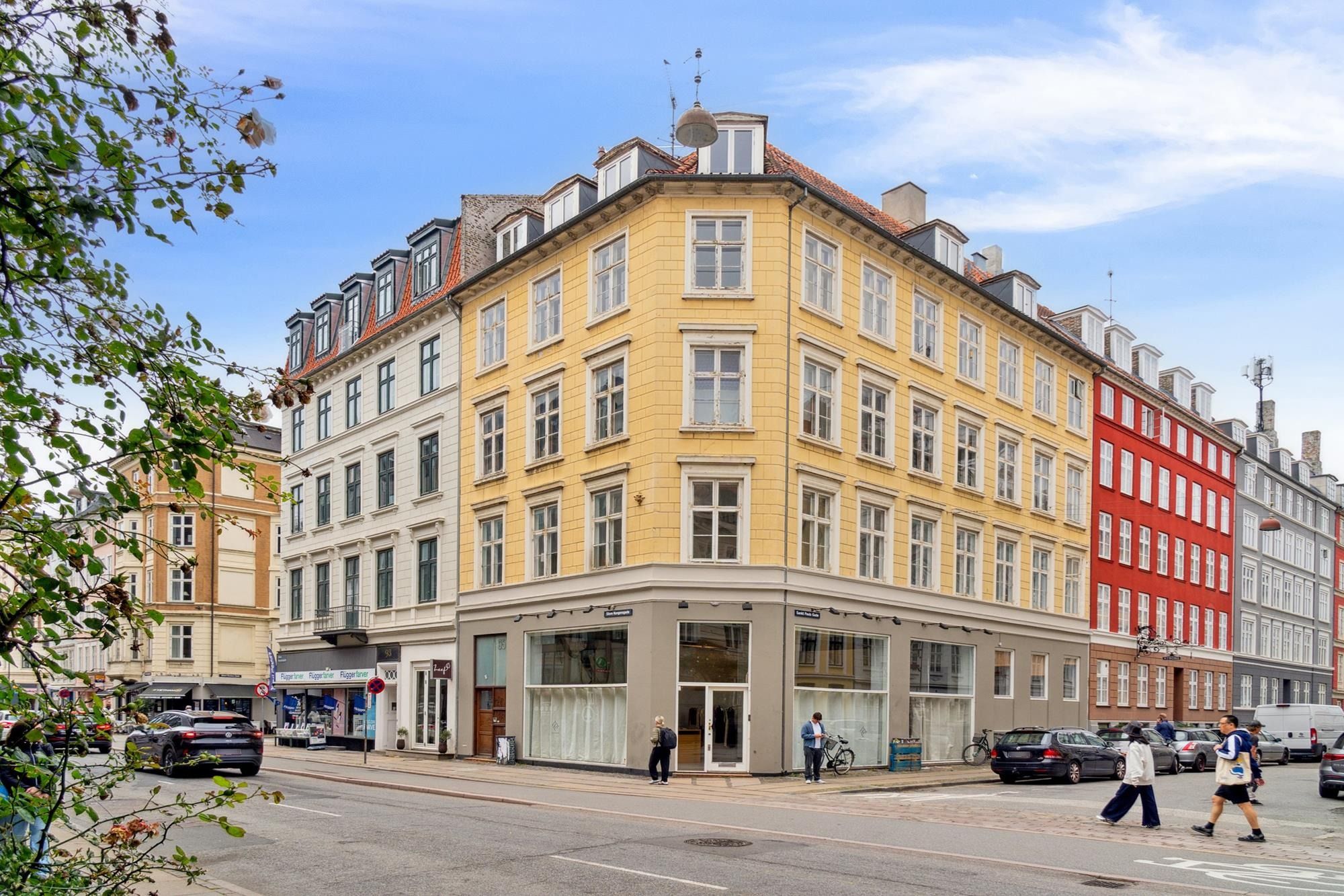 Mennesker går på gågaden med biler og cykler, røde og gule bygninger med mange vinduer, forretninger og en parkeringsplads