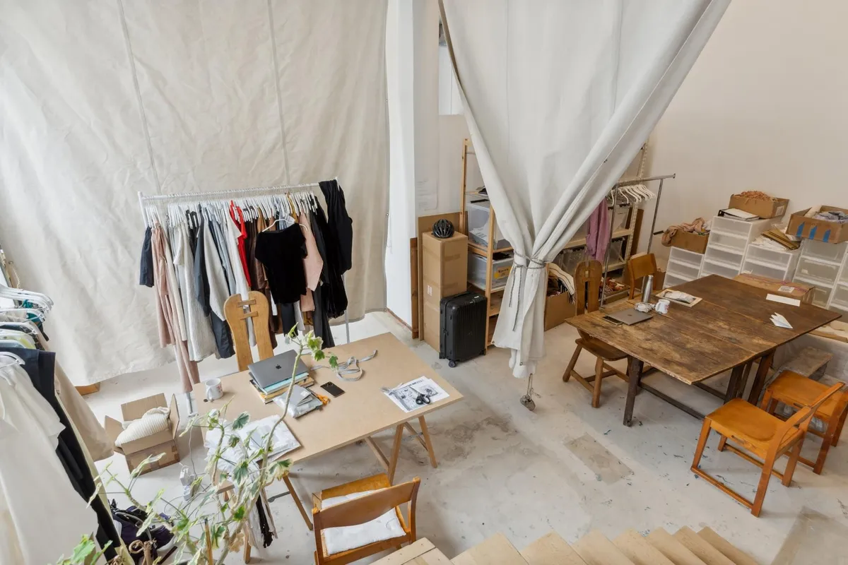 Værelse med skrivebord, stole, stol, og træbord med skråkant. Et stykke tøj i en klædeskab med tøj på et stativ. Væggen er