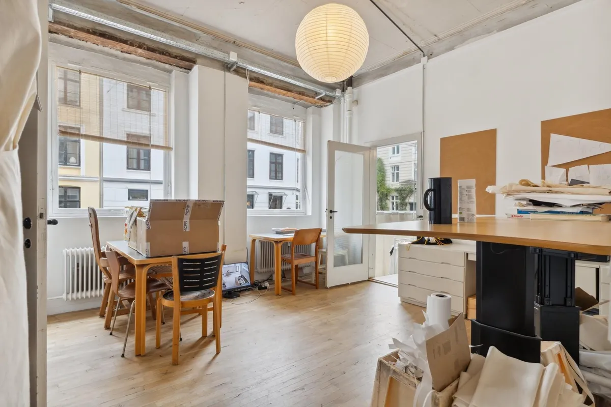 Rummelig stue med masser af plads og højt til loftet, fuld af møbler og ting, som skal pakke. Rundt lampe lyser og giver