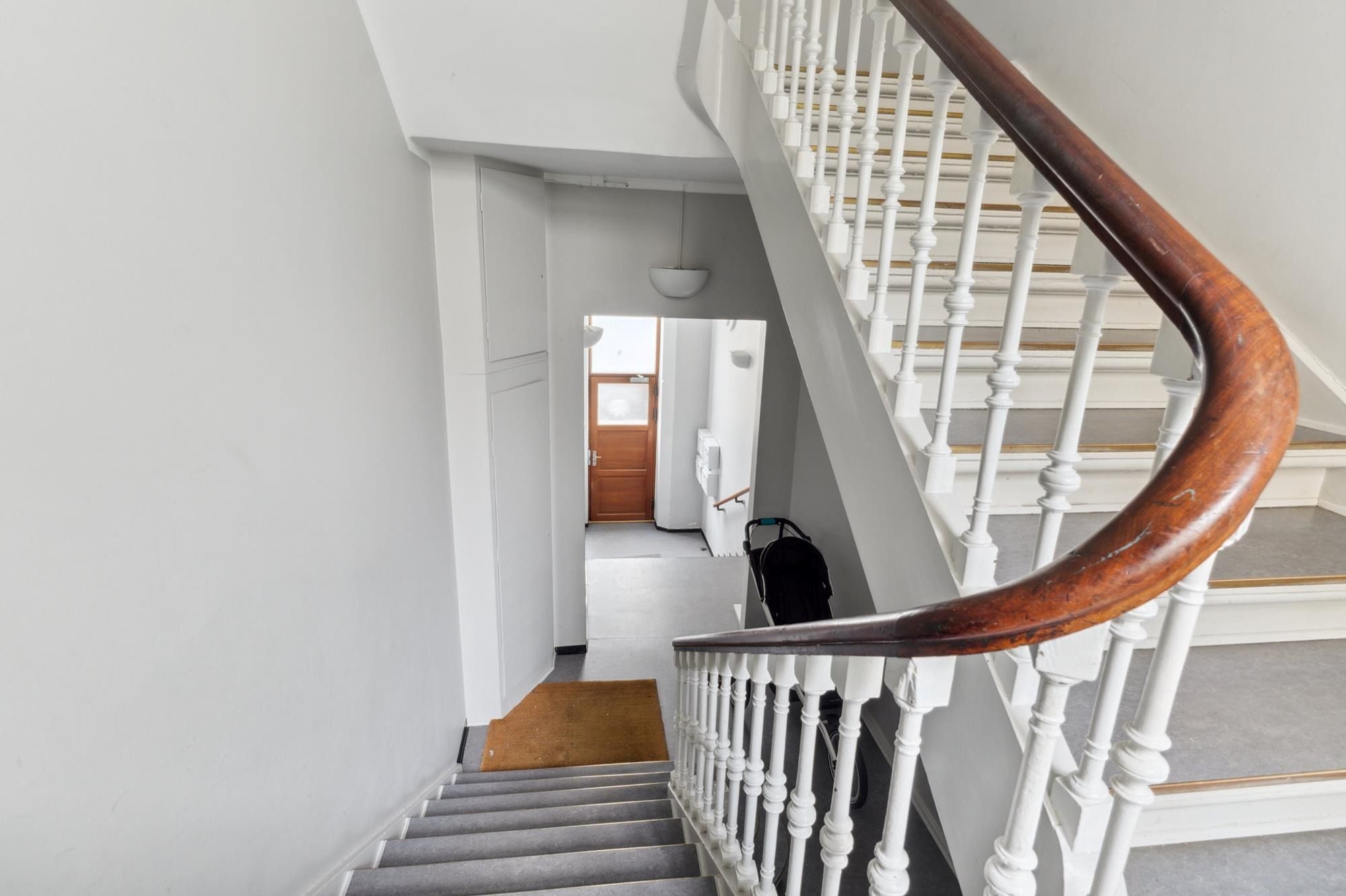 Træklatret trappe med hvidt gesims og lysegrå forløbere i en hvid interiør med trægulve, åbent træpanel og lysekroner