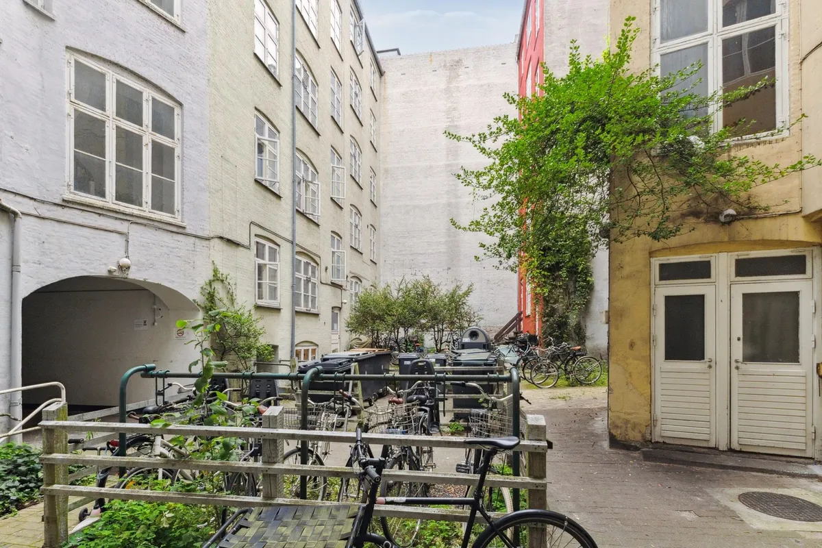 En smalle gade med to bygninger, flere parkerede cykler og et grønt træ på den ene bygning.