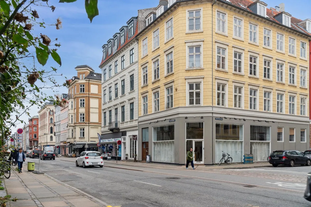 På Store Kongensgade går en person forbi butikker og køretøjer, og en cykel står parkeret ved siden af et bord med en række
