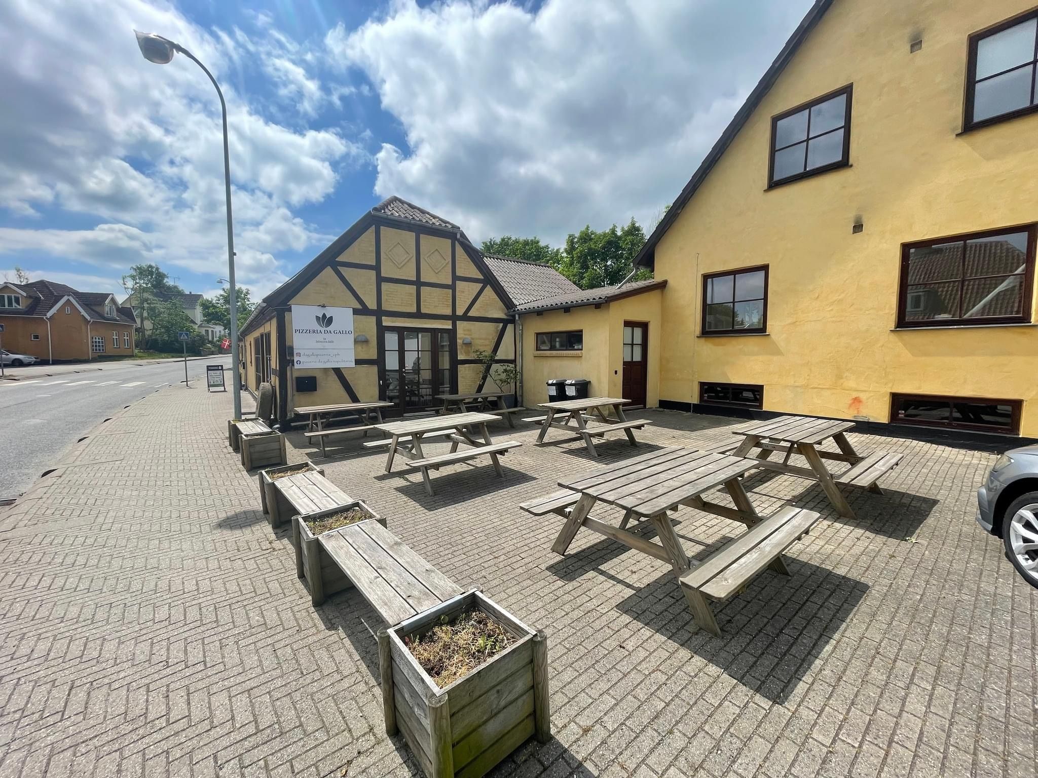 Pizzaerien med navnet Pizzaria Da Gallo og terrassen med træborde og bænke placeret foran restauranten i solskinnet