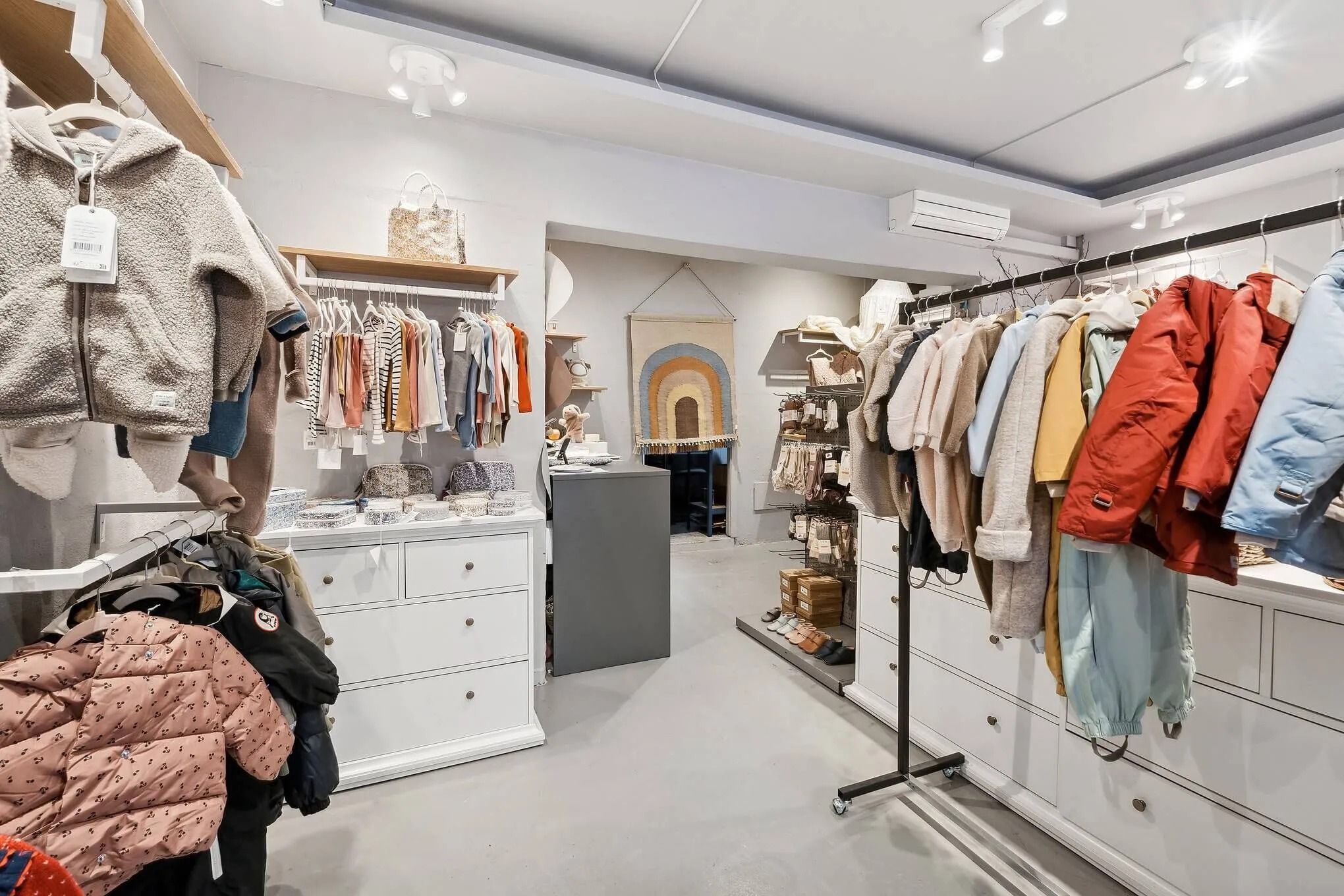 En køkkenudstilling med tøj, hættetrøjer og jakker på hylder og i skabe samt en simpel stol og et tæppe i midten af lokalet