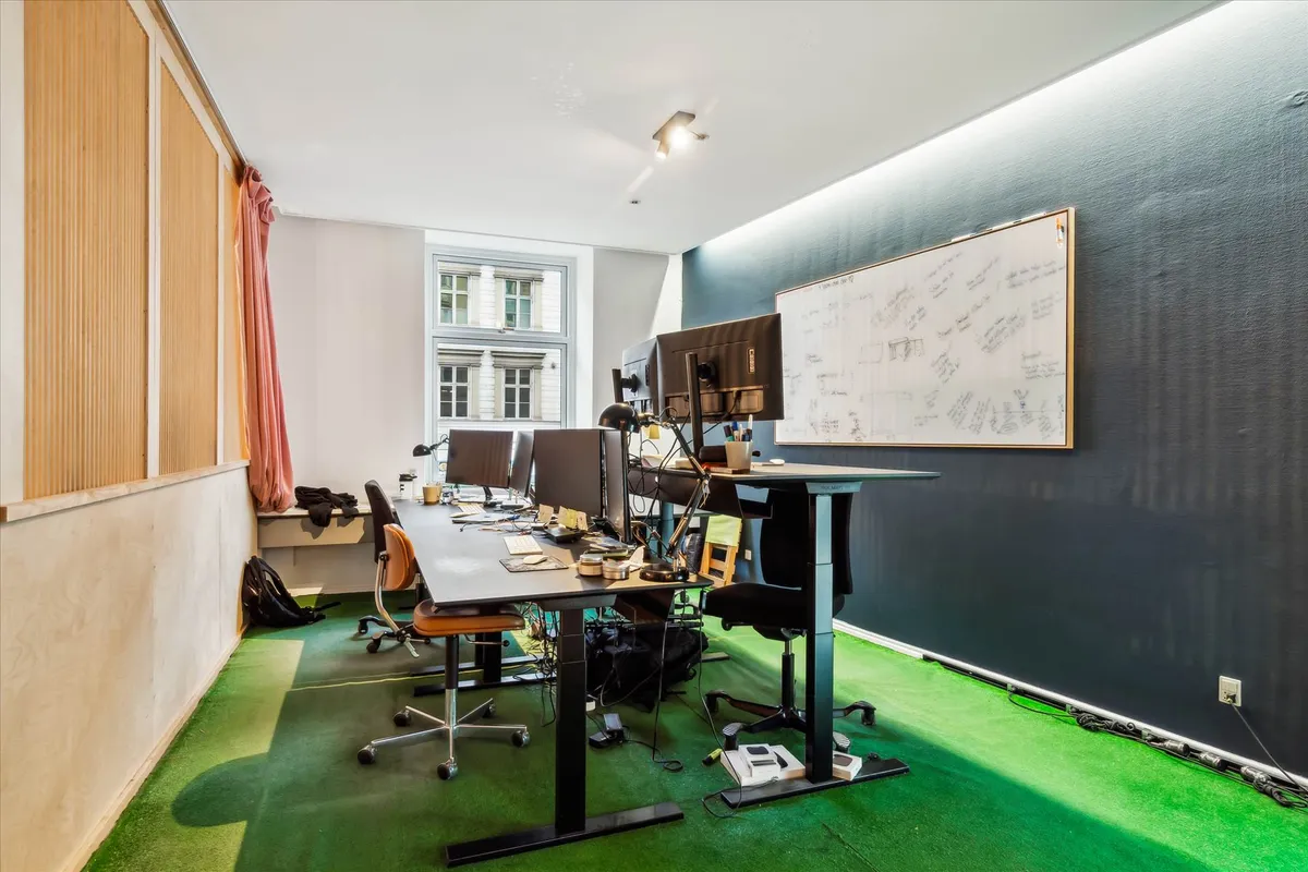 Kontorrum med flere skriveborde, computere og skrivebordslamper samt en hvidt papir med tegninger og noter på væggen.