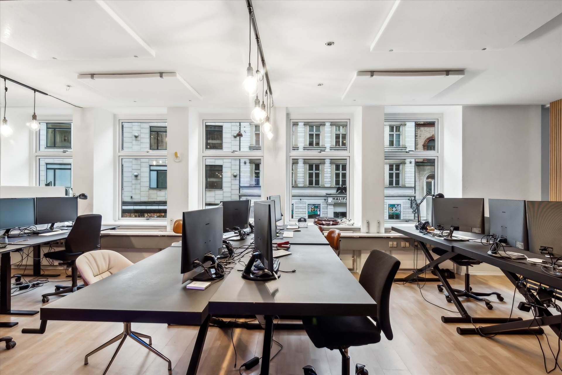 Kontor med skrivebord, stol, monitorer og slanke lysekroner. Gulve af træ og højt til loftet. Værker og vinduer giver lys.