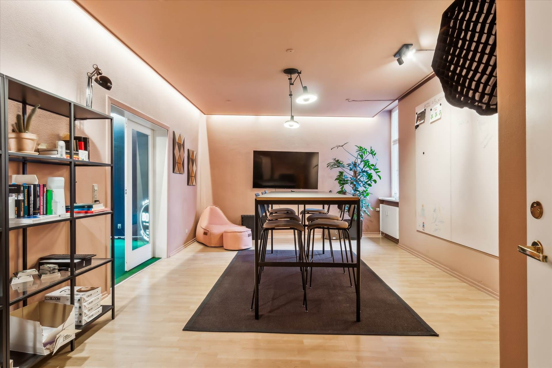 Kontor med højt til loftet, pæne trægulve og elevator. Et bord med fire stole og en sofa samt en tv-skærm. Et regnbuebord