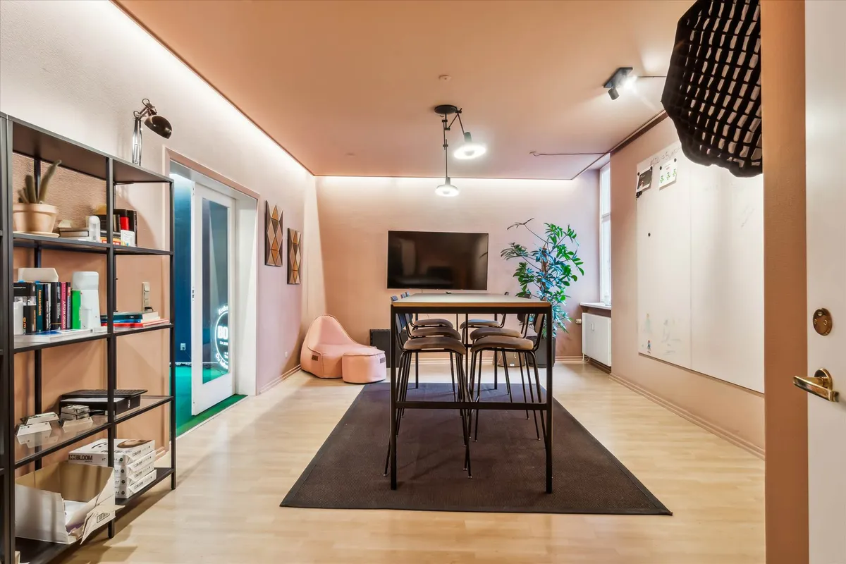 Kontor med højt til loftet, pæne trægulve og elevator. Et bord med fire stole og en sofa samt en tv-skærm. Et regnbuebord