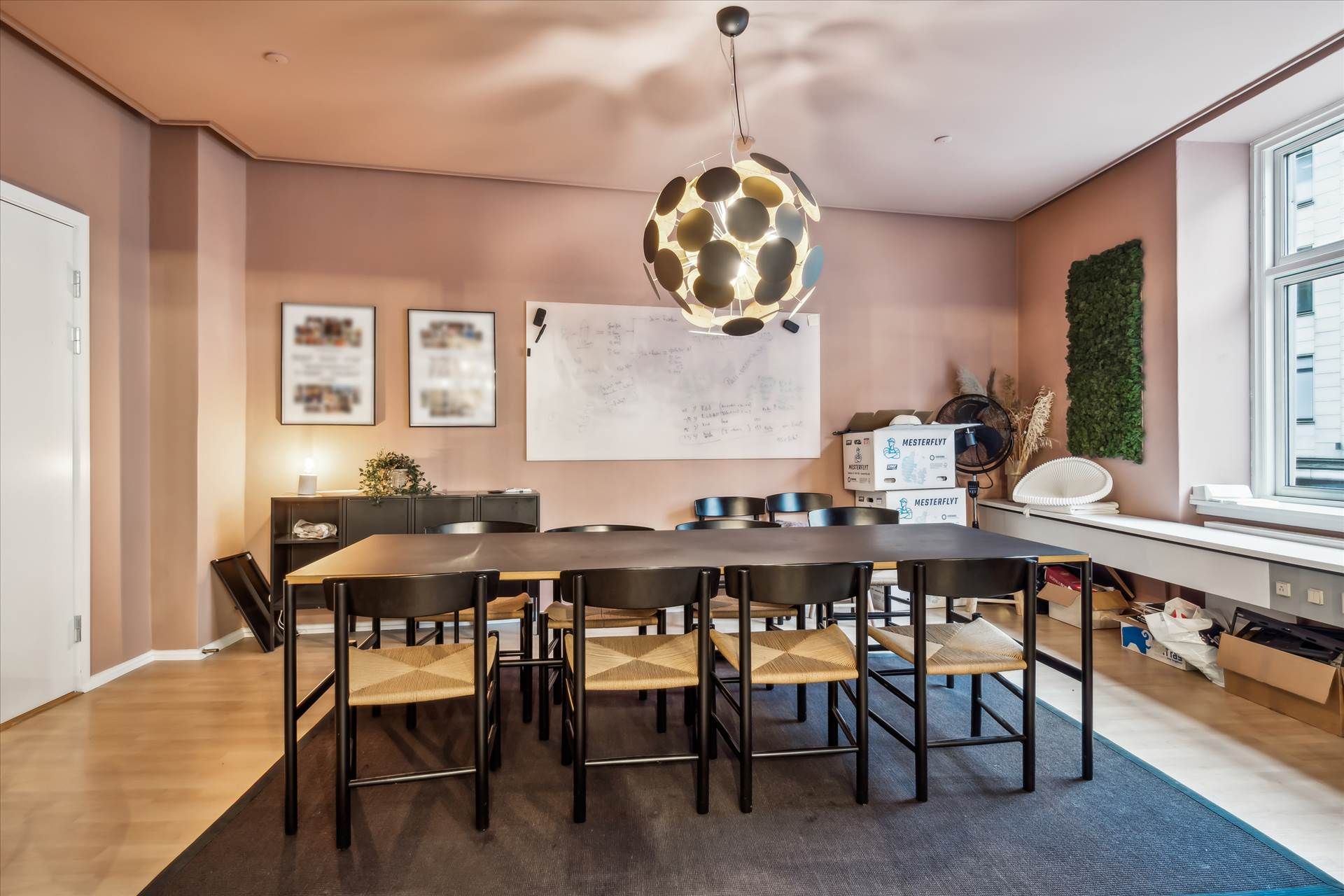 Kontor med langt spisbord og stole samt loftslampe, skrivebord og plante samt trægulv og lyserøde vægge