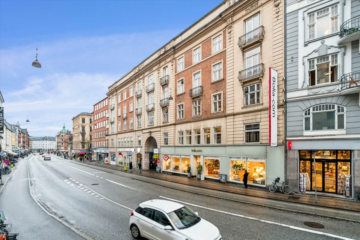 Hvid bil kører ad Værnedamsvej, med trægulve, højt til loftet og elevator i bygning. Bolig- og møbelbutikker på gadehjørnet.