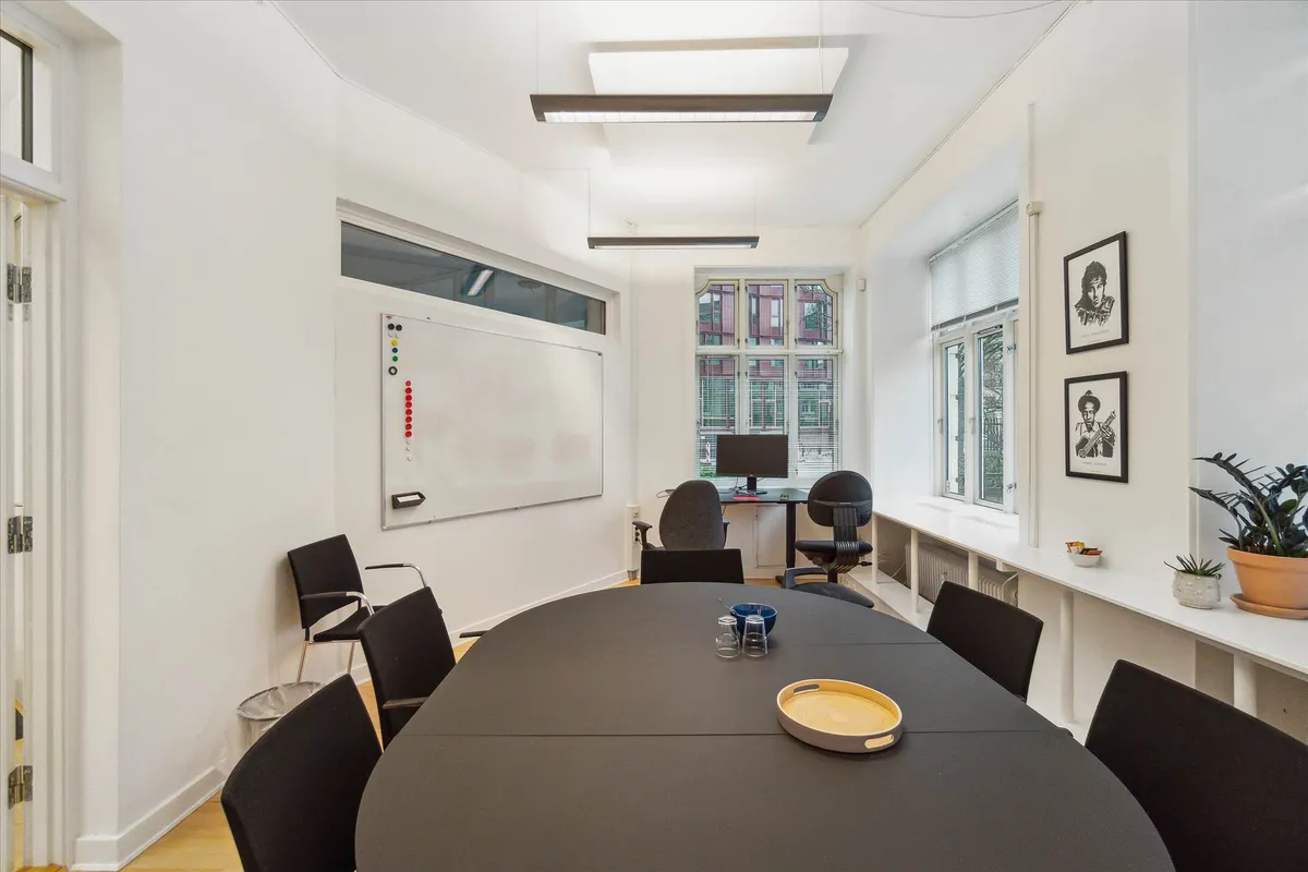 Rundt bord med to lyseborde over og to skriveborde med skærme. Vægmontører med flere portrætter og et stykke whiteboard.