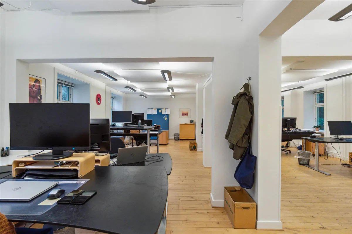 Kontor med flere skriveborde, stole, og skrivebordslamper samt computere og laptops og en brun kasse og flere klæder