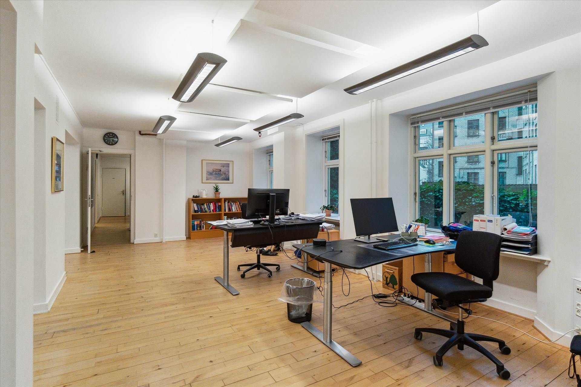 Et stort kontor med høje lofter og lyse loftslamper. Der er to store skriveborde og to skærme. Et af skrivebordene er fuldt