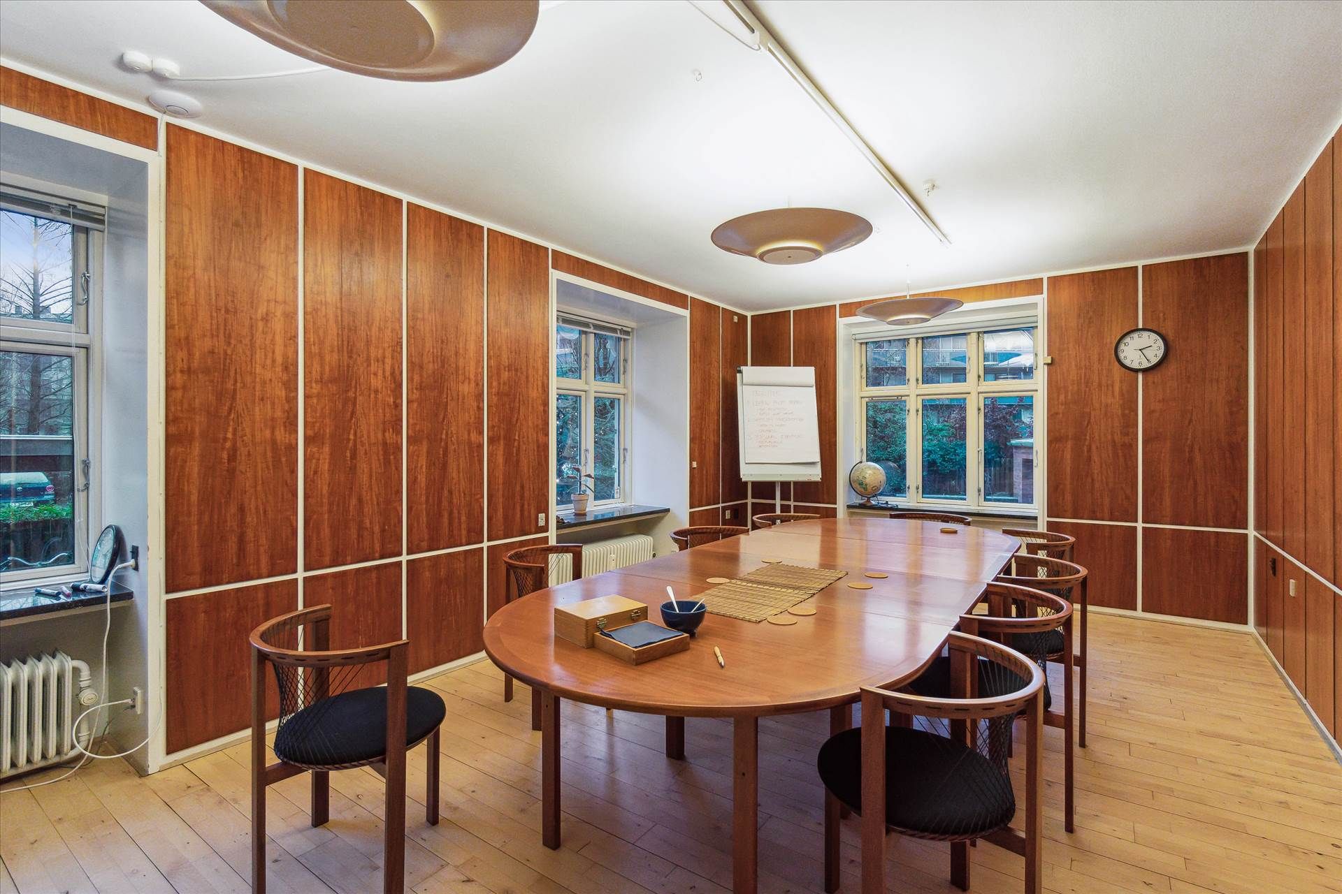 Moderne kontor med ovalt bord og stole, to hvide borde med tavle og globus, lysekroner, vindue med udsigt til gaden,