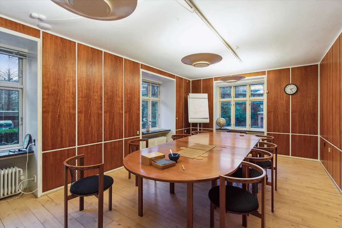 Moderne kontor med ovalt bord og stole, to hvide borde med tavle og globus, lysekroner, vindue med udsigt til gaden,