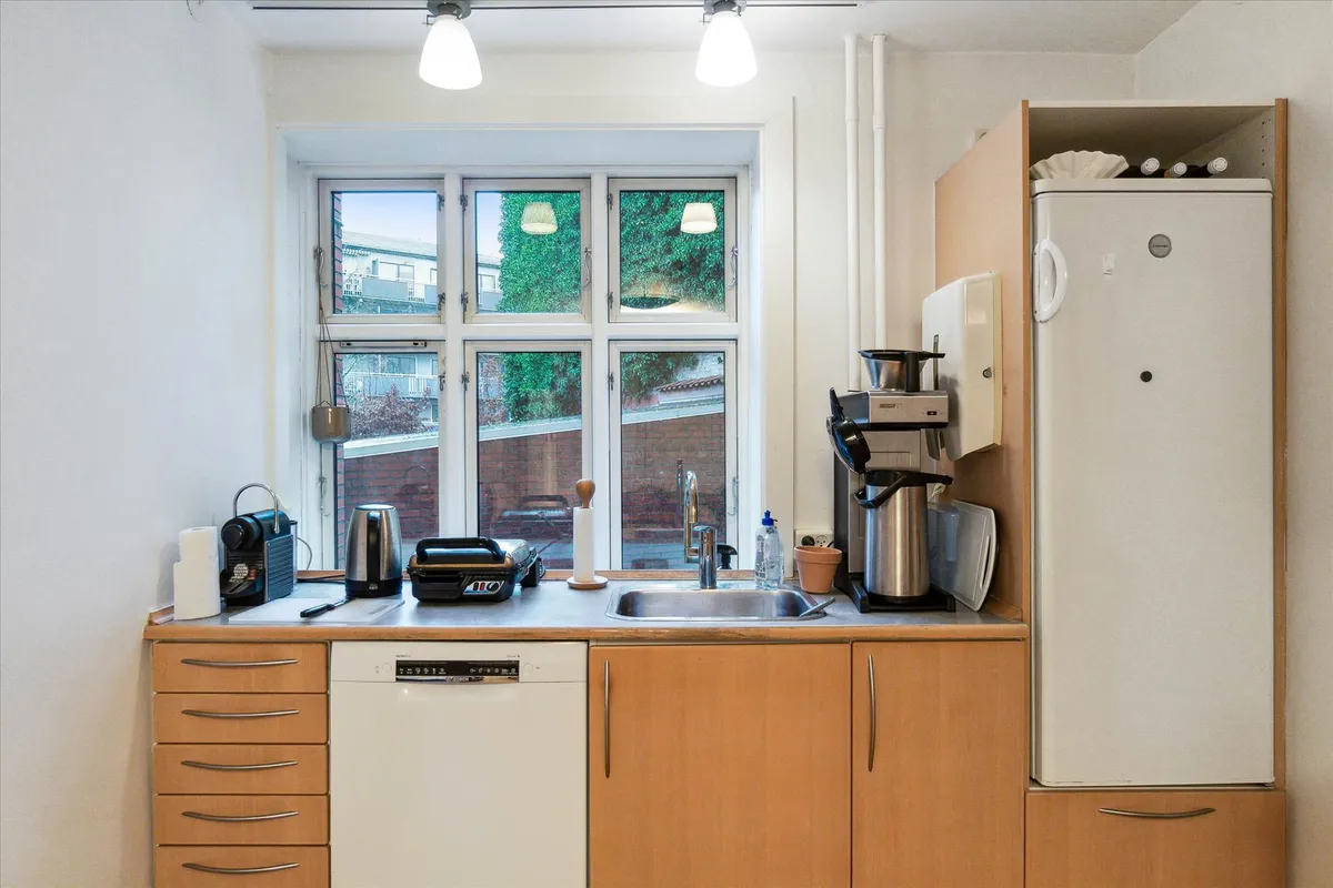 Køkken med lysegrå vægge og højt loft med to lysekroner og et vindue med udsigt til byggede huse og grønne planter