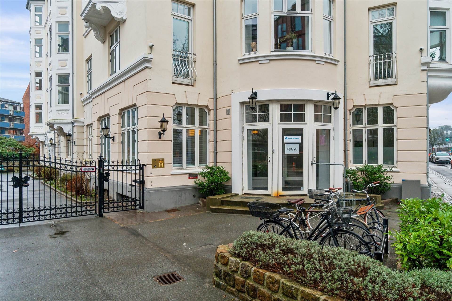 En hvidpudset bygning med fire gulve og lyseruder, udenfor er en gågade med parkerede biler og planter