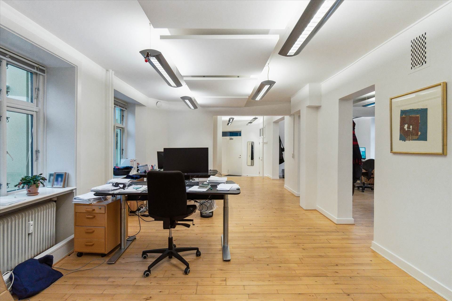 Et tomt kontor med højt til loftet, moderne møbler, en skrivebord med computer, og et lille plantepotte på vindueskarmen.