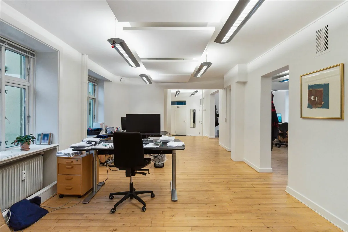 Et tomt kontor med højt til loftet, moderne møbler, en skrivebord med computer, og et lille plantepotte på vindueskarmen.