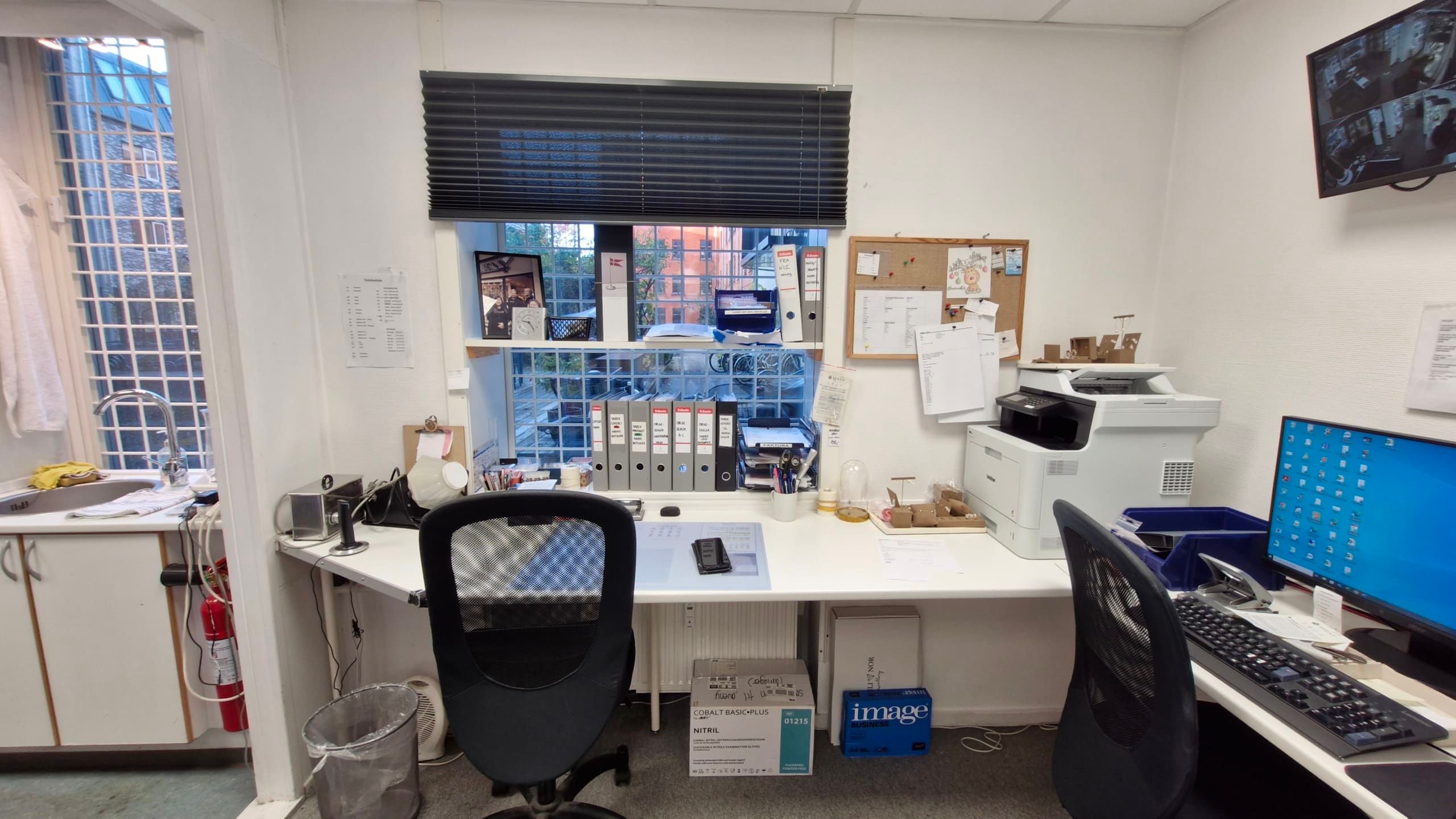 Kontor med to skriveborde og stol, printer, computer, monitor, skrivemateriale, skraldkurv, rødt brandslukker,