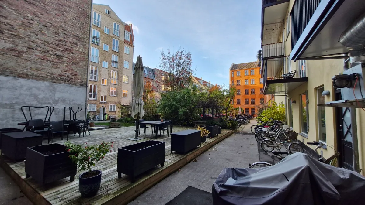 Forside med bord og stole og paraply samt potteplanter og cykler i kø