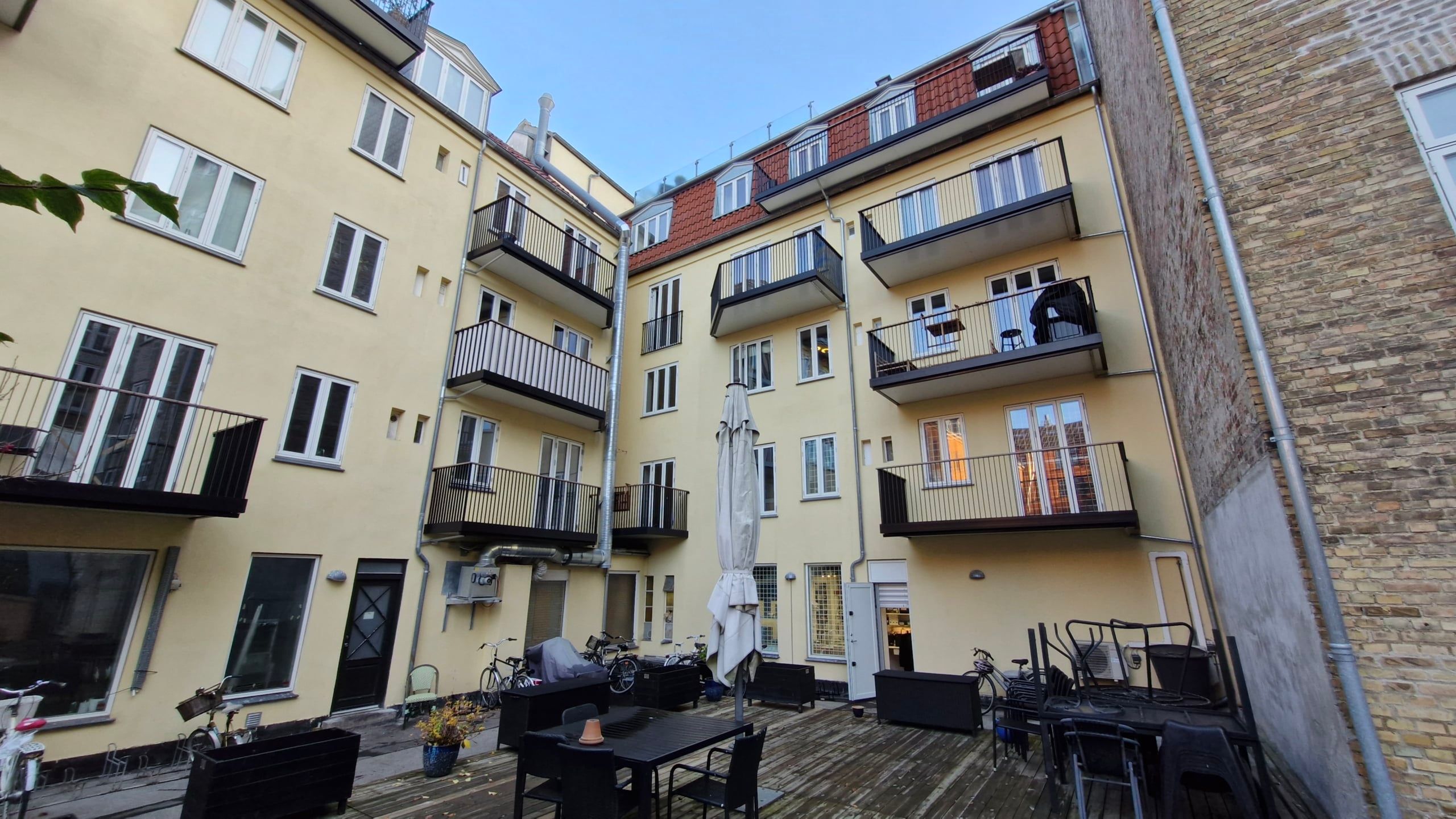 På en gård i en bygning med mange lejligheder og balkoner er der bord og stole samt en parasol