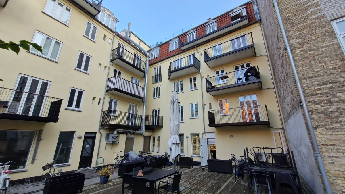 På en gård i en bygning med mange lejligheder og balkoner er der bord og stole samt en parasol
