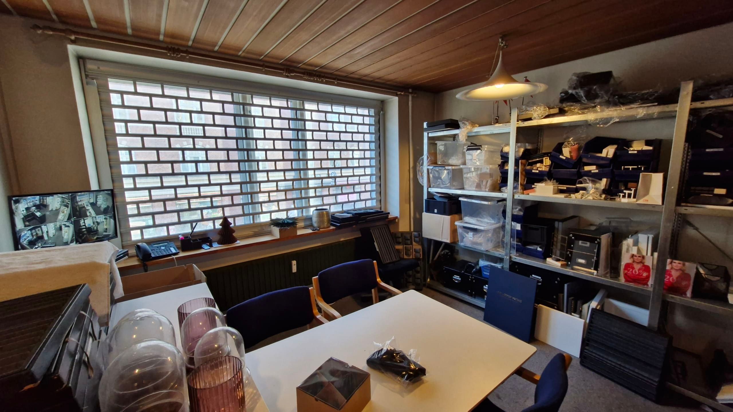 Kontorrum med skrivebord og to stolpe, opbevaringsbokse og skærme på vindueskarmen, en lampe hængende fra loftet og en