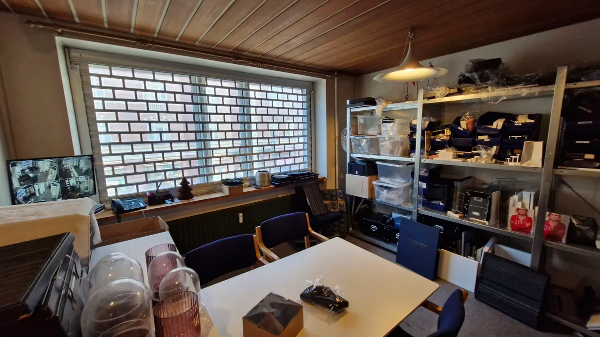 Kontorrum med skrivebord og to stolpe, opbevaringsbokse og skærme på vindueskarmen, en lampe hængende fra loftet og en